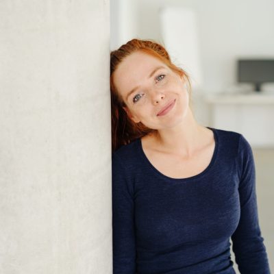 Pretty young redhead woman with a sweet sincere smile and look of kind empathy leaning against an interior pillar with copy space