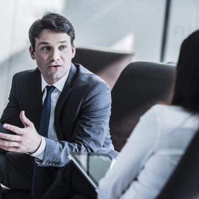 Smart mid adult business executive gesturing and talking in meeting with colleague. Serious professional man in suit at work in modern office.